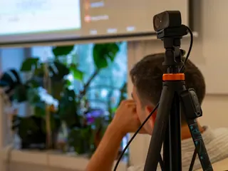 A webcam on a tripod in the foreground; behind it, a person watches the projected screen.